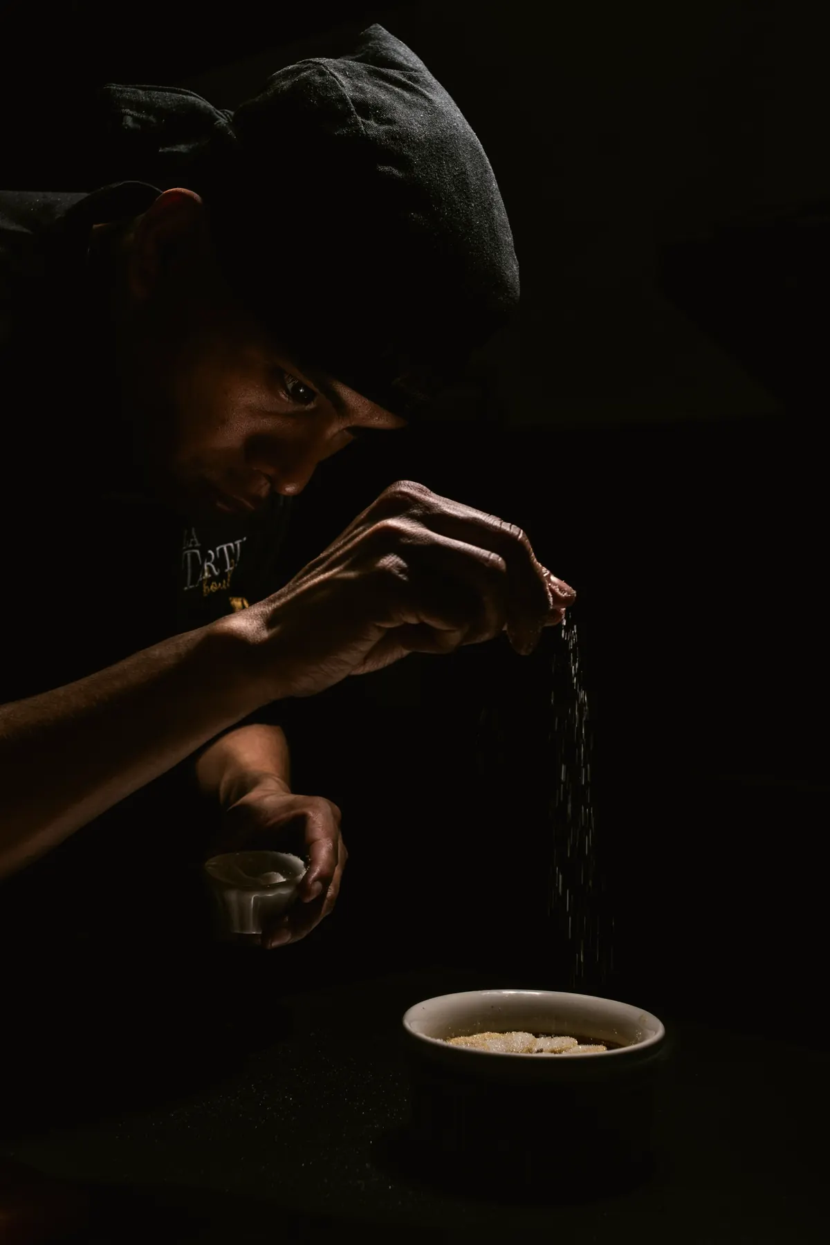 The chef, dressed in black, seasons a dish in near darkness
